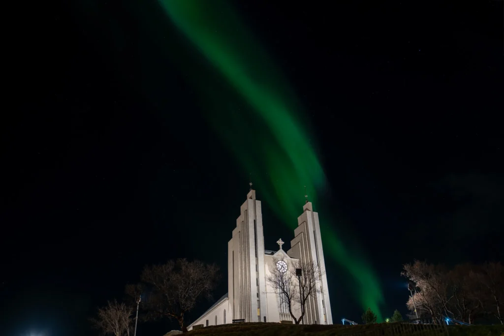 Glowing green northern lights swirling across the night sky directly above the modernist twin towers of Akureyrarkirkja church in Akureyri.