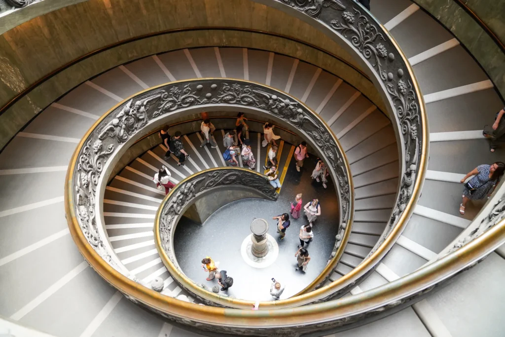 Top-down circular perspective of the ornate bronze-railed double helix Bramante spiral staircase with visitors walking down inside the Vatican Museums.