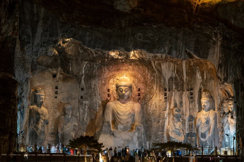 Towering rock-cut Buddhist statues including the central Vairocana Buddha and heavenly guardians illuminated in warm golden spotlights against dark limestone cliffs.