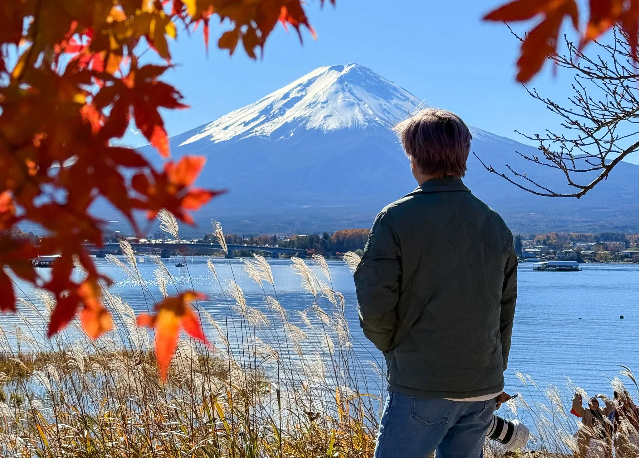 Landscape photographer with a telephoto lens looking out at snow-topped Mount Fuji across the blue lake water, framed by autumn maple leaves and reeds in Japan.