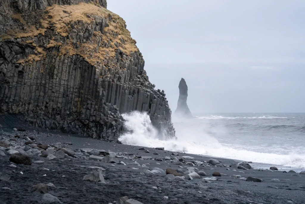 Giant hexagonal basalt rock formations on the black sand beach of Reynisfjara with wild ocean waves crashing against cliffs and the Reynisdrangar sea stack offshore.