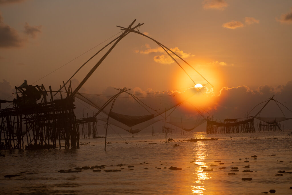 Radiant orange morning sun breaking through clouds directly behind the wooden frame of a giant traditional dipping fish net in Thale Noi, Thailand.